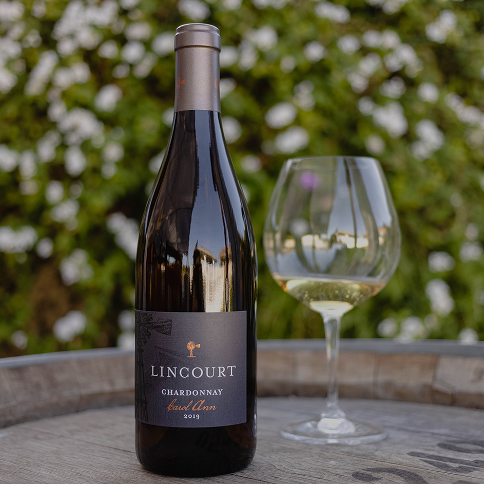 A bottle of lincourt on a table top next to a glass of white wine with a beautiful flower bush in the background